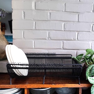 May include: A black wire dish rack holds white plates on a wooden shelf. The rack is rectangular with a sloped design. A vase with purple flowers and a potted plant are also visible.