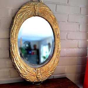 Peut inclure: Miroir ovale doré avec un cadre orné et texturé. Le cadre présente un motif de papillon en haut et en bas. Le miroir est accroché à un mur de briques grises. Une petite poignée est en haut pour l'accrocher.