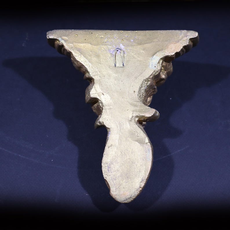May include: A gold-colored decorative wall shelf with an ornate design. The shelf has a small hole in the back for hanging.