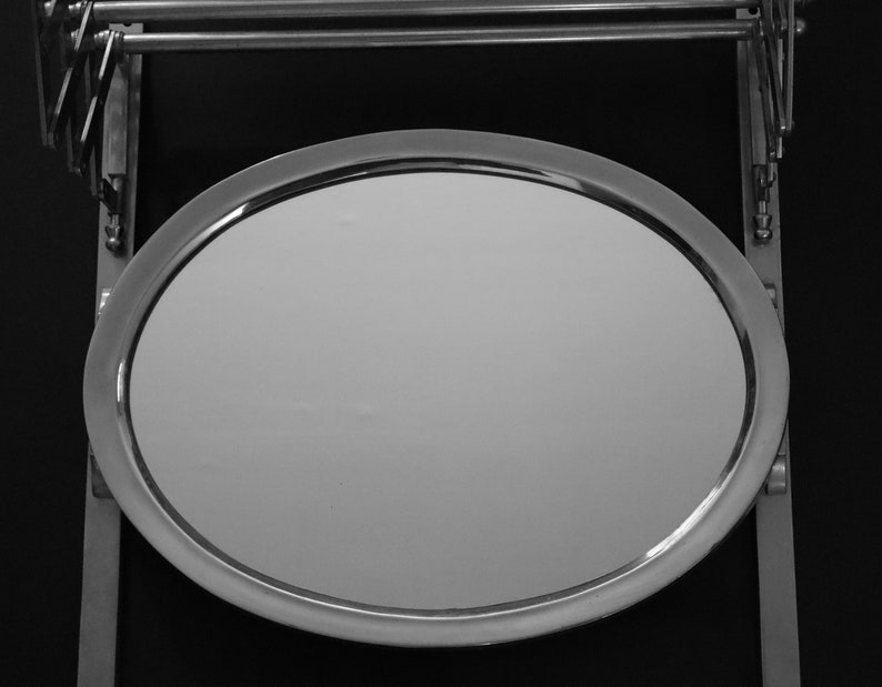 May include: A round, silver-framed mirror with a smooth, reflective surface. The mirror is attached to a metal stand with a swivel arm.