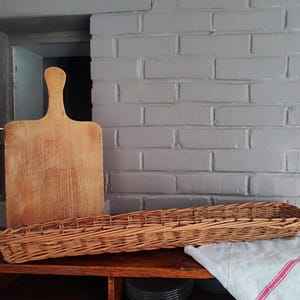 Op de afbeelding: Een lichtbruine houten snijplank met een handvat staat verticaal naast een lange, smalle, rechthoekige gevlochten rieten mand. Een witte doek met een dunne rode streep is gevouwen en gedeeltelijk onder de mand op een houten oppervlak geplaatst.