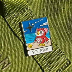 May include: A rectangular enamel pin featuring a red owl character with a star and ocean background. The pin has a white border and the words "THE STAR" at the bottom. The pin is set against a green fabric background.