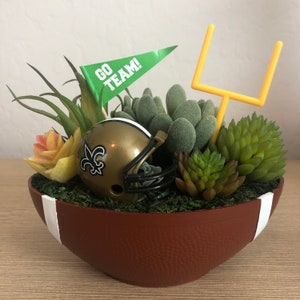 May include: A football-shaped planter with a brown exterior and white stripes. It contains a miniature gold football helmet, a green flag that says "GO TEAM!", a yellow goalpost, and various succulents and greenery.