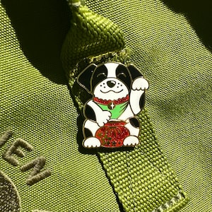 May include: A colorful enamel pin featuring a lucky dog design. The pin has a white body with black ears and spots, a green shirt, and a red collar. The dog is holding a red and gold money bag. The pin is attached to a green fabric strap.