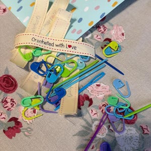 May include: A selection of crochet tools, including stitch markers and needles, are displayed on a floral patterned surface. A ribbon with the text "Crocheted with Love" is also present. The tools are in shades of blue, green, and purple.