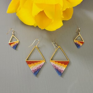 May include: A set of three pairs of gold-toned earrings with a geometric diamond shape. The earrings are beaded with a rainbow of colors, including red, orange, yellow, green, blue, indigo, and violet.