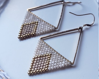 May include: A pair of gold triangle earrings with white and beige beaded fringe. The earrings are hanging from a white cord with gold accents.