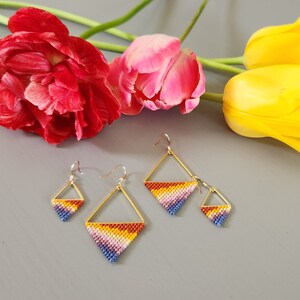 May include: Three pairs of geometric, rainbow-colored beaded earrings with gold wire accents. The earrings are arranged on a gray surface with yellow and pink tulips in the background.