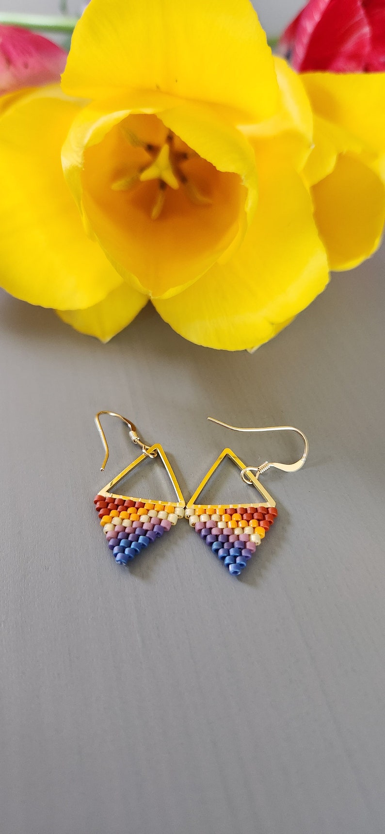 May include: A pair of triangle-shaped earrings with a rainbow color pattern. The earrings are made of small beads and have silver hooks.
