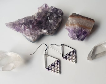 May include: A pair of silver dangle earrings with a geometric design. The earrings are made with small purple and white beads. The earrings are displayed on a white surface with amethyst and clear quartz crystals.
