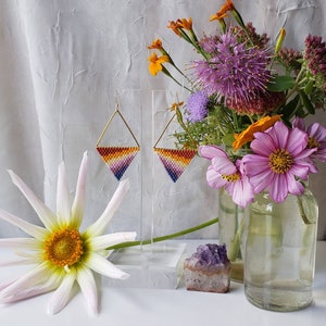 May include: A pair of triangle-shaped earrings with a gradient of orange, yellow, pink, and purple beads. The earrings are hanging from a gold-colored wire and are displayed on a white stand. The earrings are in front of a bouquet of flowers in a glass vase.