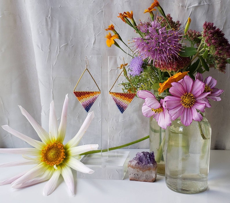 May include: A pair of triangle-shaped earrings with a gradient of orange, yellow, pink, and purple beads. The earrings are hanging from a gold-colored wire and are displayed on a white stand. The earrings are in front of a bouquet of flowers in a glass vase.