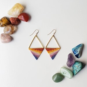 May include: A pair of gold triangle earrings with a rainbow gradient of beads. The earrings are on a white background with colorful stones.