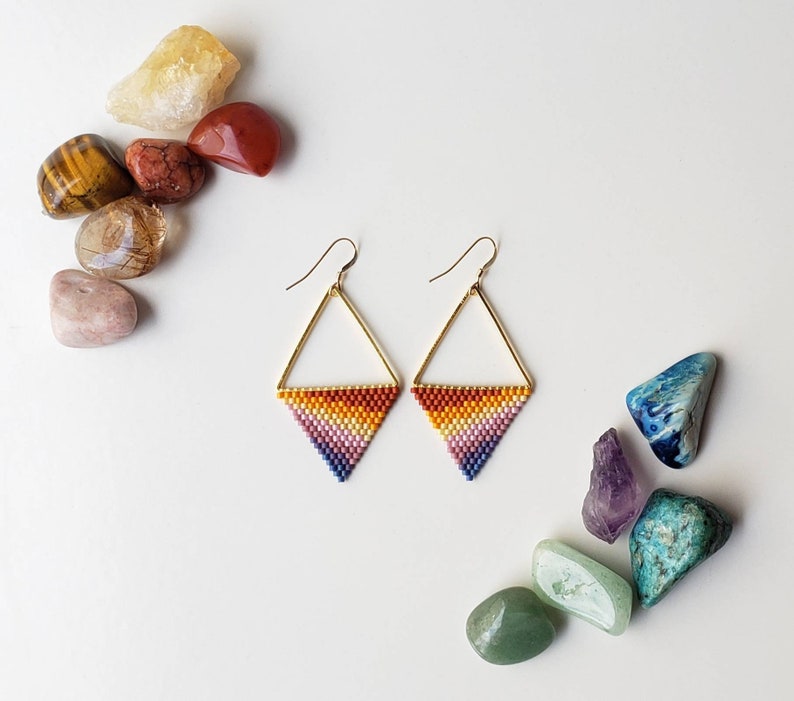 May include: A pair of gold triangle earrings with a rainbow gradient of beads. The earrings are on a white background with colorful stones.