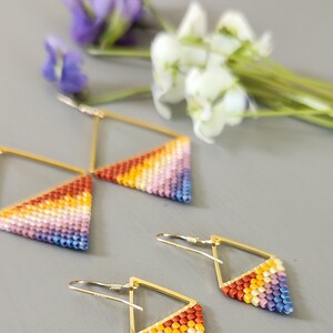 May include: A pair of gold triangle earrings with a rainbow gradient of beads. The earrings are made with small, colorful beads and have a geometric shape.