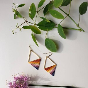 May include: A pair of gold triangle earrings with a rainbow gradient of beads. The earrings are on a white surface with green leaves and a purple flower.