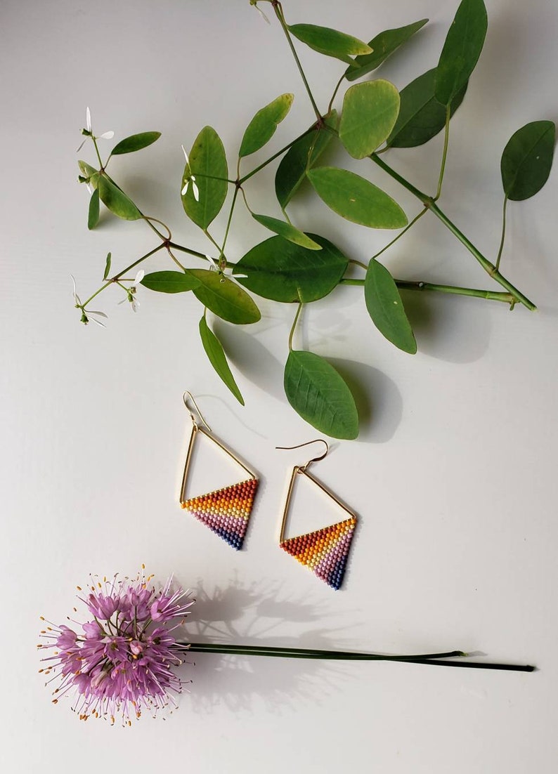 May include: A pair of gold triangle earrings with a rainbow gradient of beads. The earrings are on a white surface with green leaves and a purple flower.