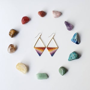 May include: A pair of gold triangle earrings with rainbow colored beads. The earrings are surrounded by various colored stones.