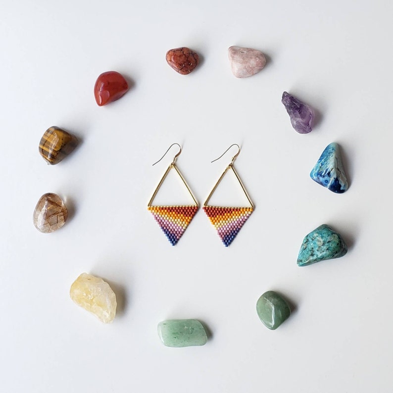 May include: A pair of gold triangle earrings with rainbow colored beads. The earrings are surrounded by various colored stones.