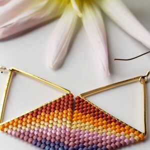 May include: A pair of gold triangle earrings with a multi-colored beaded fringe. The fringe is made of small, square beads in shades of orange, yellow, pink, and purple.