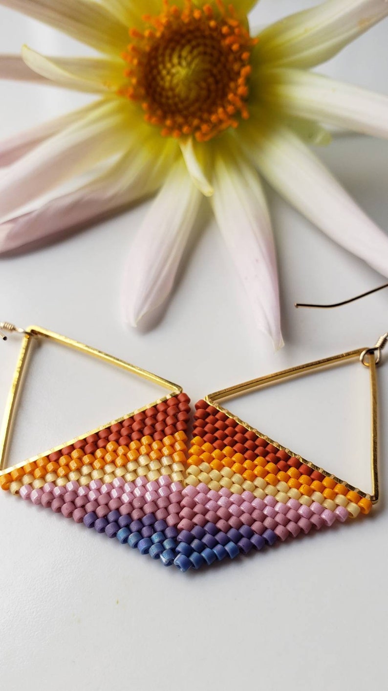 May include: A pair of gold triangle earrings with a multi-colored beaded fringe. The fringe is made of small, square beads in shades of orange, yellow, pink, and purple.