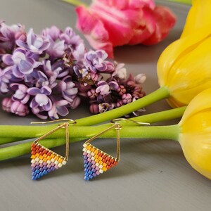 May include: A pair of rainbow-colored beaded triangle earrings with gold wire and hooks. The earrings are displayed on a gray surface with yellow and pink flowers.