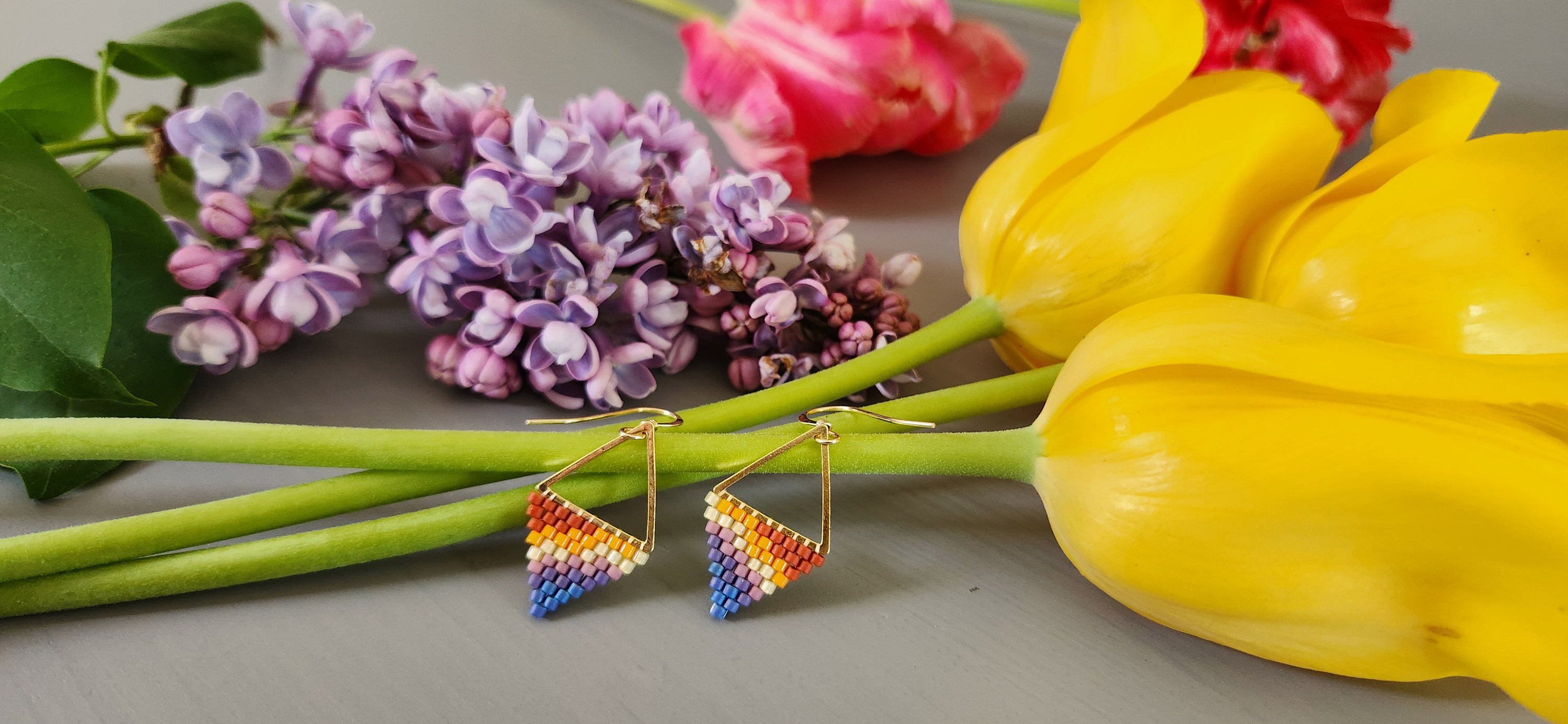May include: A pair of rainbow-colored beaded triangle earrings with gold wire and hooks. The earrings are displayed on a gray surface with yellow and pink flowers.