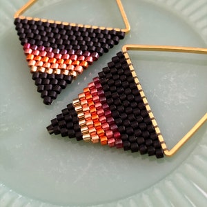 May include: A pair of gold triangle earrings with black and orange beaded fringe. The earrings are on a light green surface.