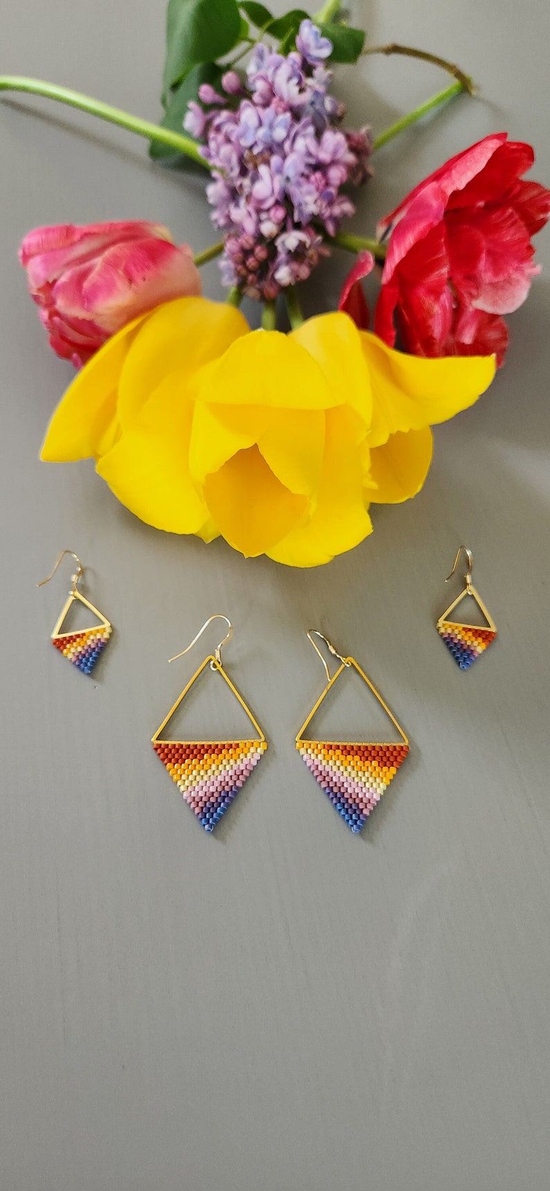 May include: A set of three pairs of gold-toned earrings with a geometric diamond shape. The earrings are beaded with a rainbow of colors, including red, orange, yellow, green, blue, indigo, and violet.