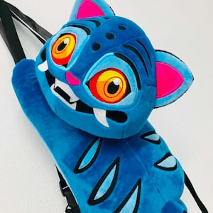 May include: A blue plush tiger-shaped backpack with orange and yellow eyes, pink ears, and white fangs. It features black stripes and black straps. The backpack is designed to be worn on the back.