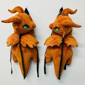 May include: Two plush dragon toys in a warm orange hue, each with green eyes and detailed wings. The dragons have curved horns and are suspended against a white background. Black straps hang from the bottom of each dragon.