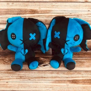 May include: Two plush bunny dolls with a split black and blue design. Each bunny has button eyes, an embroidered mouth, and a stitched cross on one cheek. A broken heart is stitched on the chest. The bunnies are on a wooden surface.