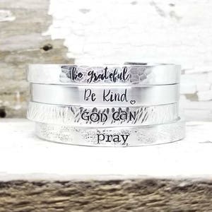 May include: Four silver bracelets stacked on top of each other. The bracelets are engraved with the words "be grateful", "be kind", "God can", and "pray".