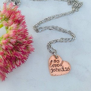 May include: A silver chain necklace with a copper heart pendant. The heart pendant has the words "so loved john 3:16" engraved on it.