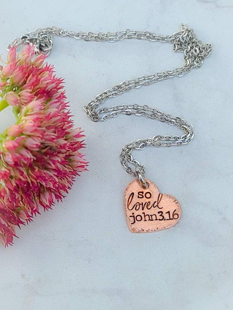 May include: A silver chain necklace with a copper heart pendant. The heart pendant has the words "so loved john 3:16" engraved on it.