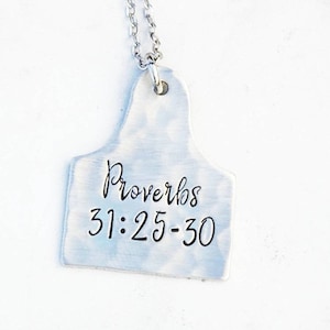 Puede incluir: Collar de plata con un colgante de etiqueta de plata que dice "Proverbios 31:25-30" en letras negras.