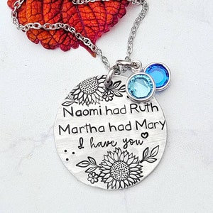 Może przedstawiać: Srebrny naszyjnik z okrągłą zawieszką z napisem "Naomi had Ruth, Martha had Mary, I have you". Zawieszka ozdobiona jest motywami słonecznik&oacute;w. Dwa niebieskie kryształowe zawieszki zwisają z łańcuszka.