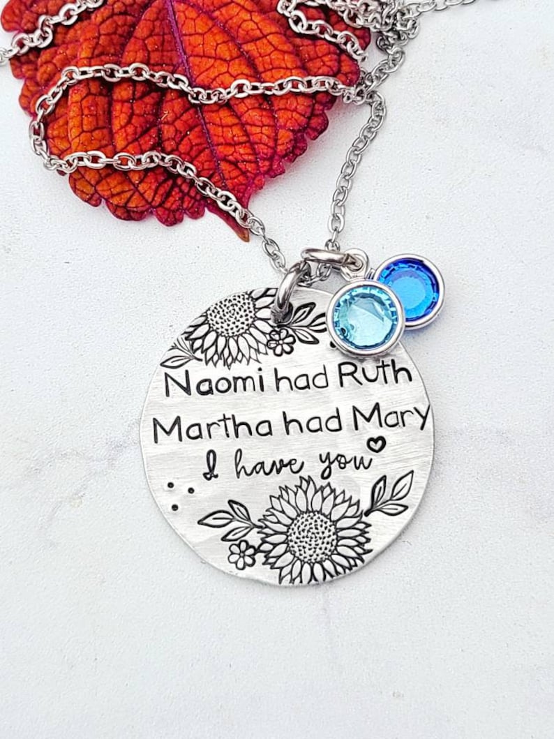 Może przedstawiać: Srebrny naszyjnik z okrągłą zawieszką z napisem "Naomi had Ruth, Martha had Mary, I have you". Zawieszka ozdobiona jest motywami słonecznik&oacute;w. Dwa niebieskie kryształowe zawieszki zwisają z łańcuszka.