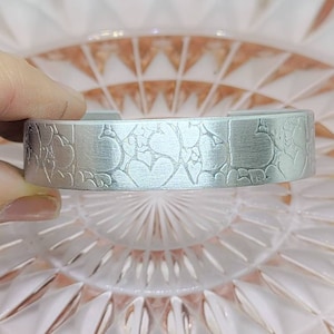 May include: A silver bracelet with a textured design of hearts and swirls. The bracelet is on a pink background.