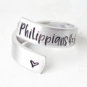 Puede incluir: Un anillo abierto de metal plateado con acabado cepillado. La banda tiene grabado el texto negro "Philippians 4:6" en la parte superior y un peque&ntilde;o contorno de coraz&oacute;n negro simple en el extremo inferior que se superpone.