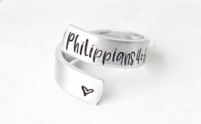 Puede incluir: Un anillo abierto de metal plateado con acabado cepillado. La banda tiene grabado el texto negro "Philippians 4:6" en la parte superior y un peque&ntilde;o contorno de coraz&oacute;n negro simple en el extremo inferior que se superpone.