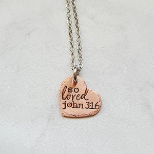 May include: A silver chain necklace with a copper heart pendant. The heart has the words "so loved john 316" engraved on it.