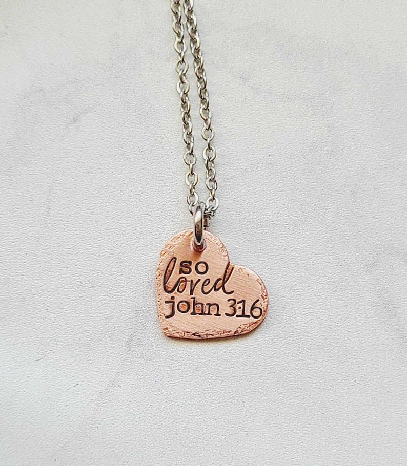 May include: A silver chain necklace with a copper heart pendant. The heart has the words "so loved john 316" engraved on it.