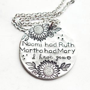 Może przedstawiać: Srebrny naszyjnik z wisiorkiem. Okrągła zawieszka ma wygrawerowany tekst i kwiatowe wzory. Tekst brzmi "Naomi miała Rut, Marta miała Marię, ja mam ciebie". Zawieszka ma wzory słonecznik&oacute;w i liści.