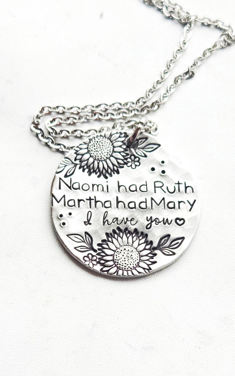 Może przedstawiać: Srebrny naszyjnik z wisiorkiem. Okrągła zawieszka ma wygrawerowany tekst i kwiatowe wzory. Tekst brzmi "Naomi miała Rut, Marta miała Marię, ja mam ciebie". Zawieszka ma wzory słonecznik&oacute;w i liści.