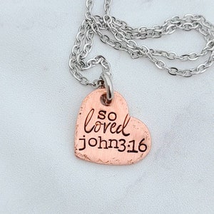 May include: A copper heart-shaped pendant necklace with a silver chain. The pendant has the words "so loved john 3:16" engraved on it.