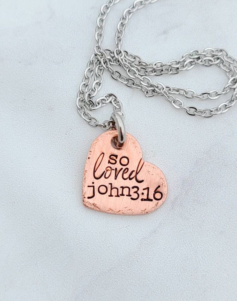 May include: A copper heart-shaped pendant necklace with a silver chain. The pendant has the words "so loved john 3:16" engraved on it.
