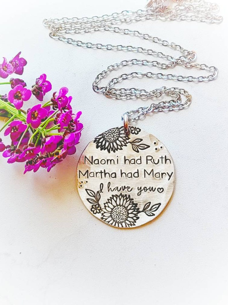 Może przedstawiać: Srebrny naszyjnik z okrągłą zawieszką. Zawieszka ma wygrawerowany tekst: &bdquo;Naomi miała Rut, Marta miała Marię, ja mam ciebie&rdquo;. Wzory kwiatowe i liści otaczają tekst. Bukiet fioletowych kwiat&oacute;w znajduje się w tle.