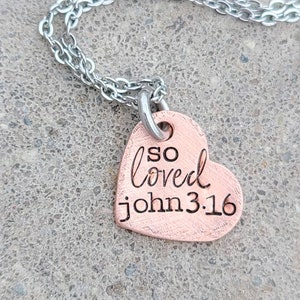 May include: A copper heart-shaped pendant necklace with a silver chain. The pendant has the words "so loved John 3:16" engraved on it.