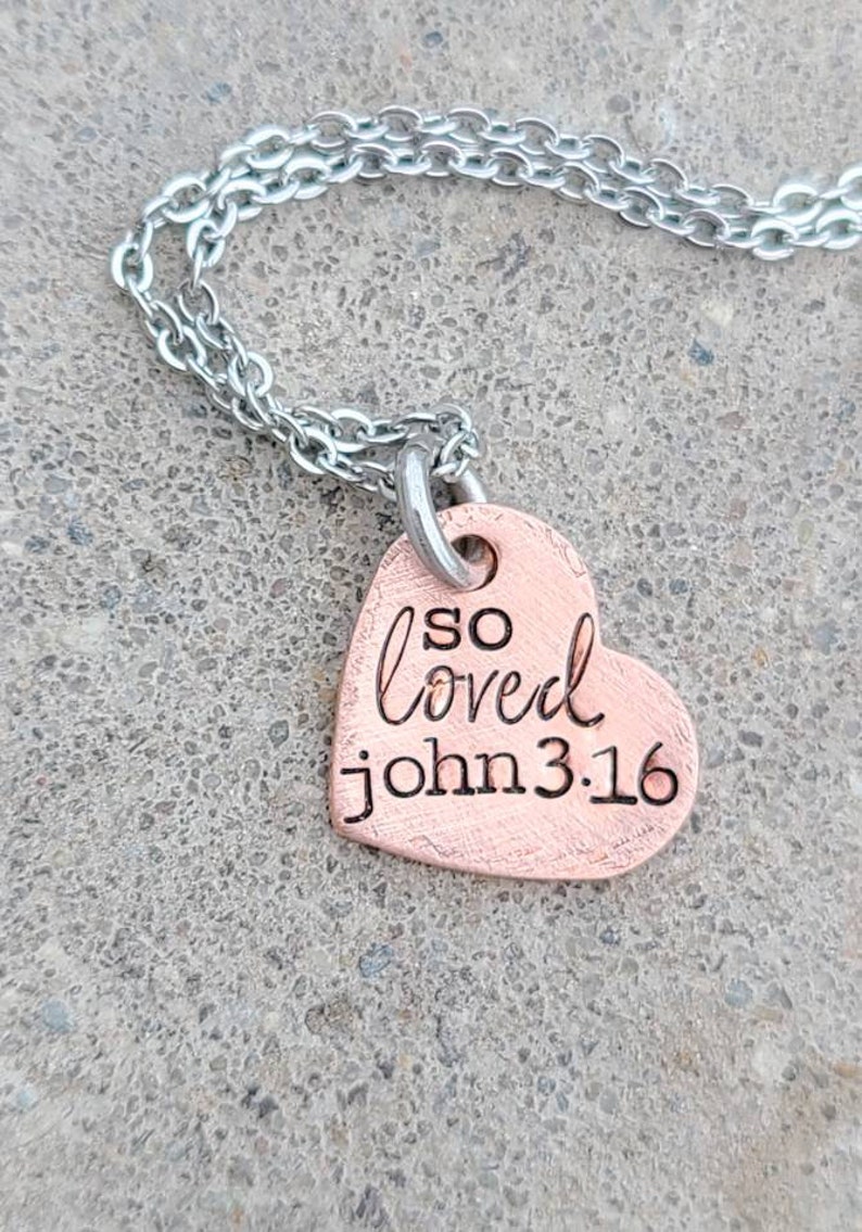 May include: A copper heart-shaped pendant necklace with a silver chain. The pendant has the words "so loved John 3:16" engraved on it.